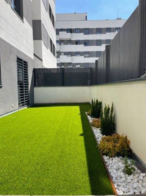 Pequeño patio con césped de color verde brillante, paredes blancas y una franja de rocas con arbustos a un lado.