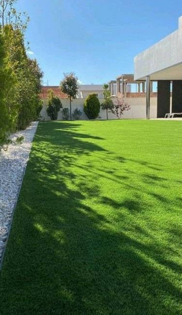 Patio urbano estrecho con césped verde brillante, paredes blancas y un pequeño parterre con piedras blancas.