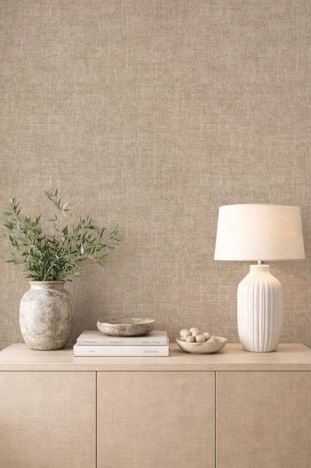 Consola minimalista de color beige con jarrón, libros, cuencos y una lámpara de mesa encendida contra una pared texturizada.