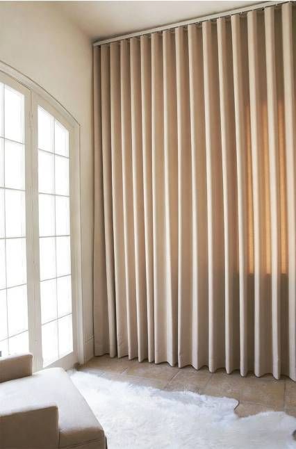 Habitación luminosa con cortinas altas de color beige, ventanas arqueadas y una alfombra blanca en el suelo.