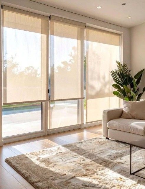 Luminoso salón con persianas translúcidas, sofá beige, alfombra y planta en maceta junto a grandes ventanales.