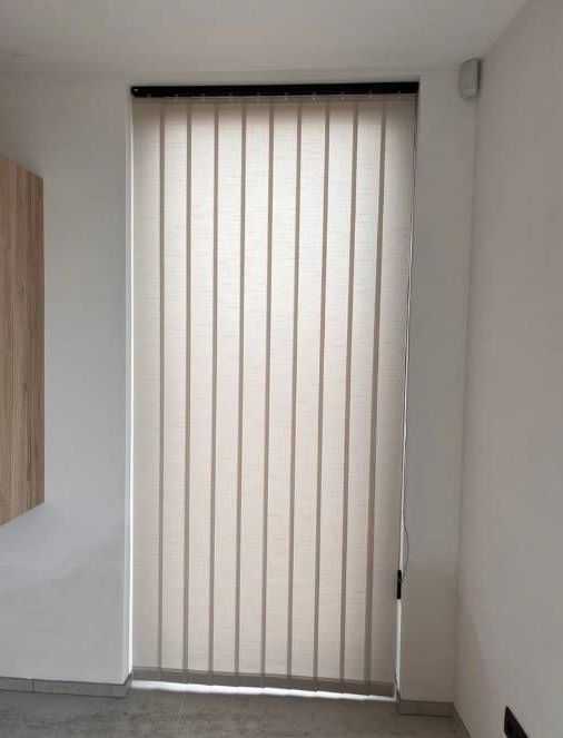 Persianas verticales beige cerradas que cubren una puerta de cristal en un pasillo blanco.