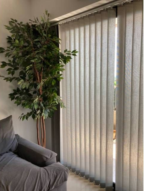 Sala de estar con una silla gris, una planta alta en maceta y persianas verticales que cubren una puerta corrediza de vidrio.