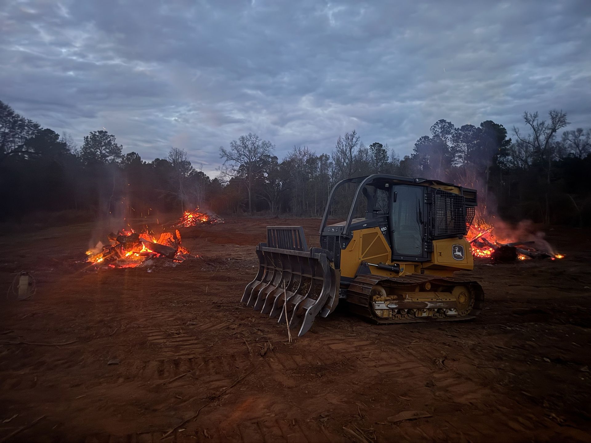 Yellow bulldozer clearing land with brush fires under a cloudy sky.