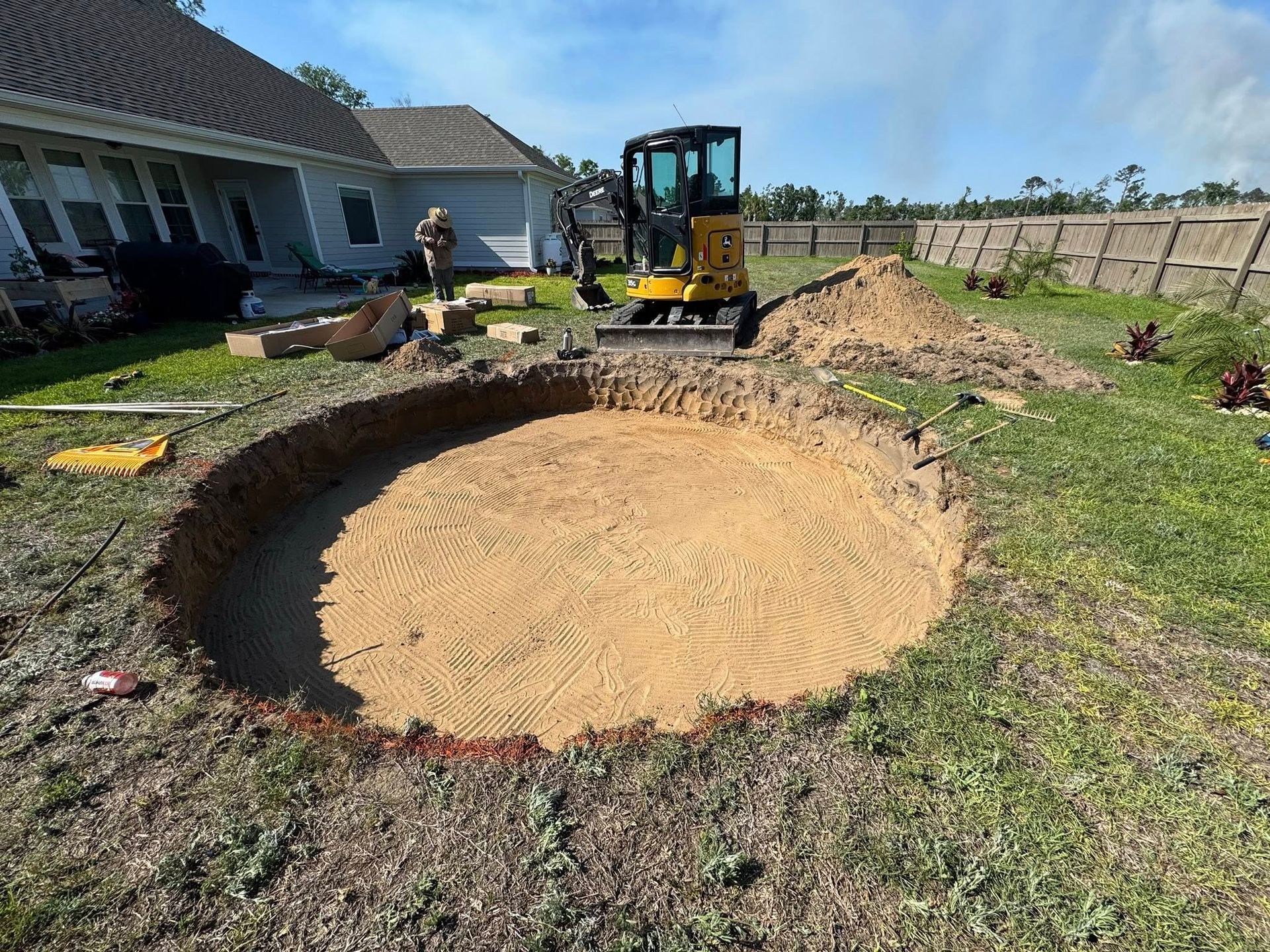 Excavation of a round area in a backyard, yellow excavator, dirt pile, and person.