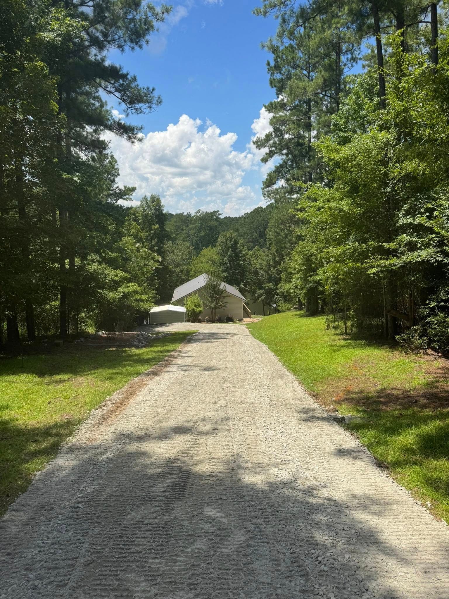 Gravel driveway leads to a light-colored house surrounded by trees on a sunny day.