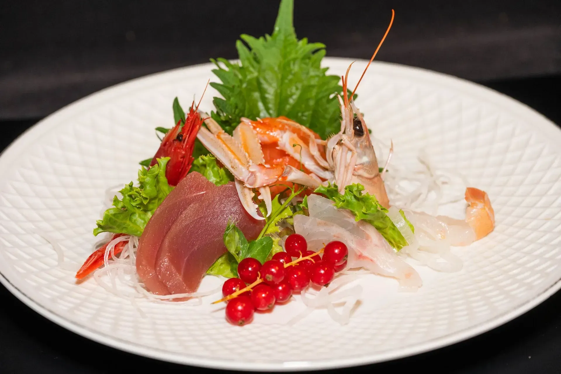 Sashimi assortito su un piatto bianco, guarnito con erbe aromatiche e ribes rosso.