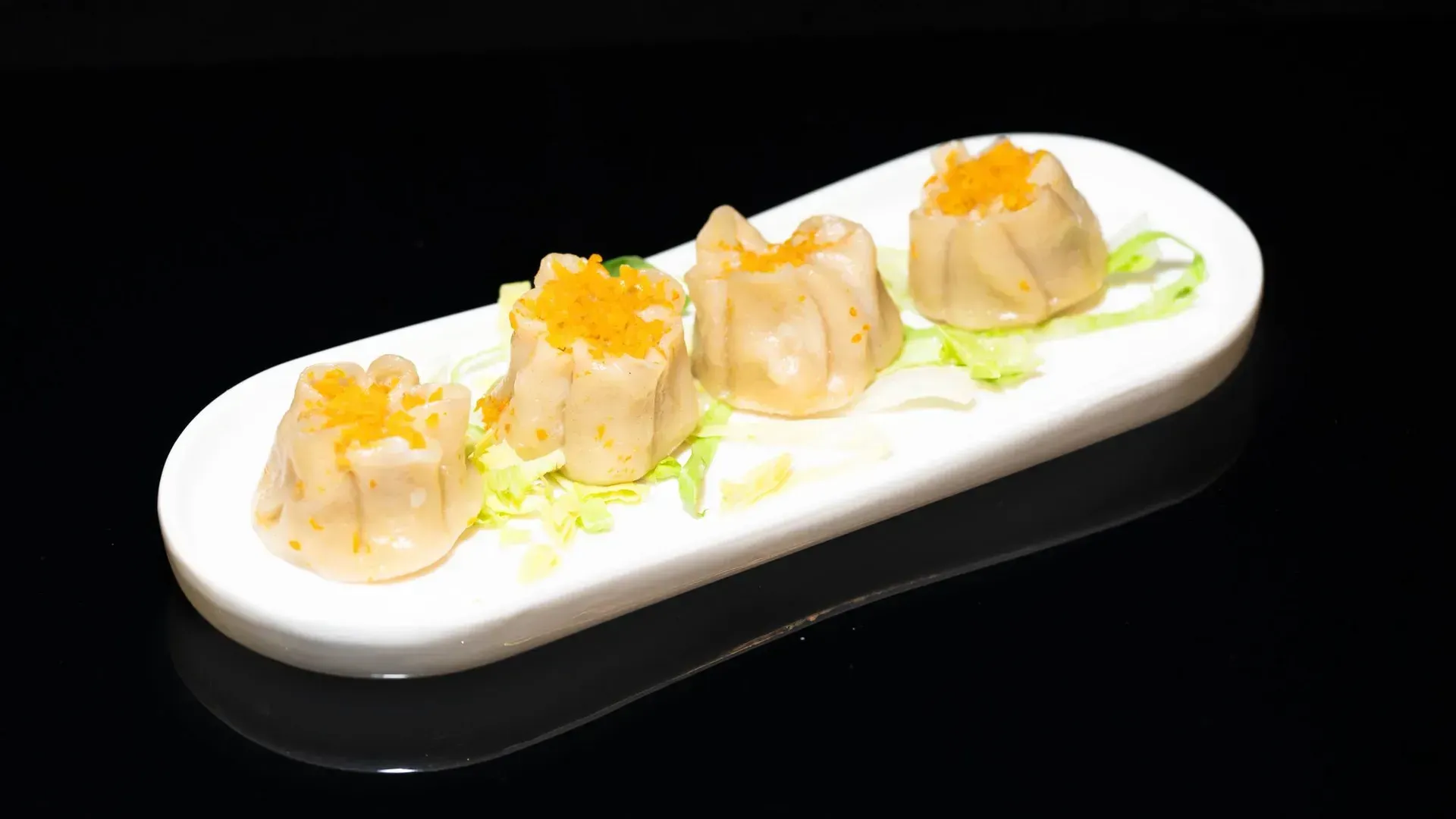 Quattro ravioli al vapore su un piatto rettangolare bianco con guarnizione verde e condimento arancione.