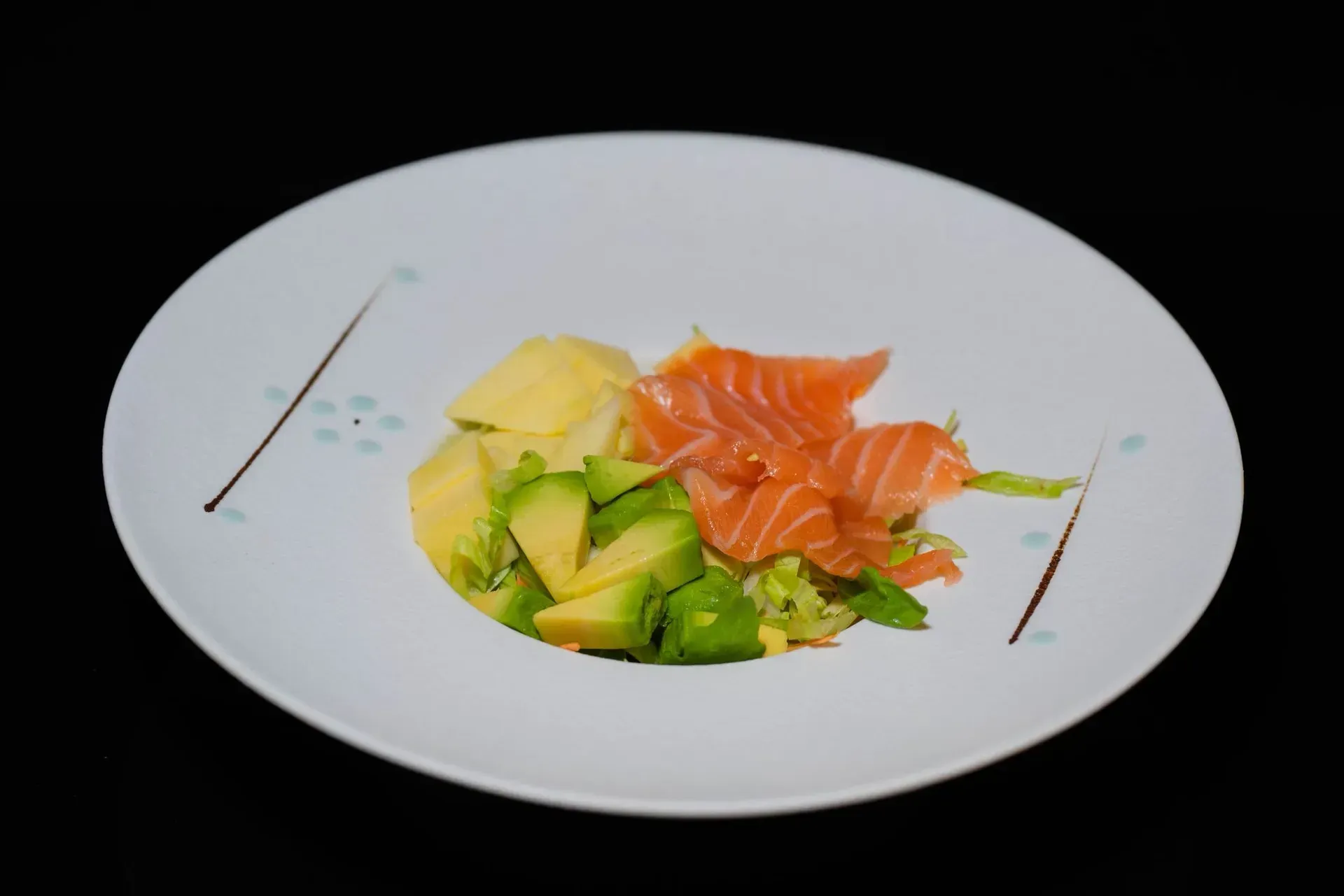 Piatto bianco con sashimi di salmone, avocado e guarnizione su sfondo nero.