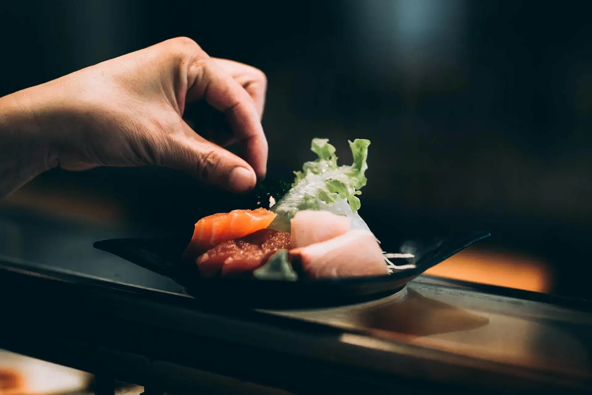Una mano che dispone il cibo su un piatto nero con guarnizione in un ristorante scarsamente illuminato.