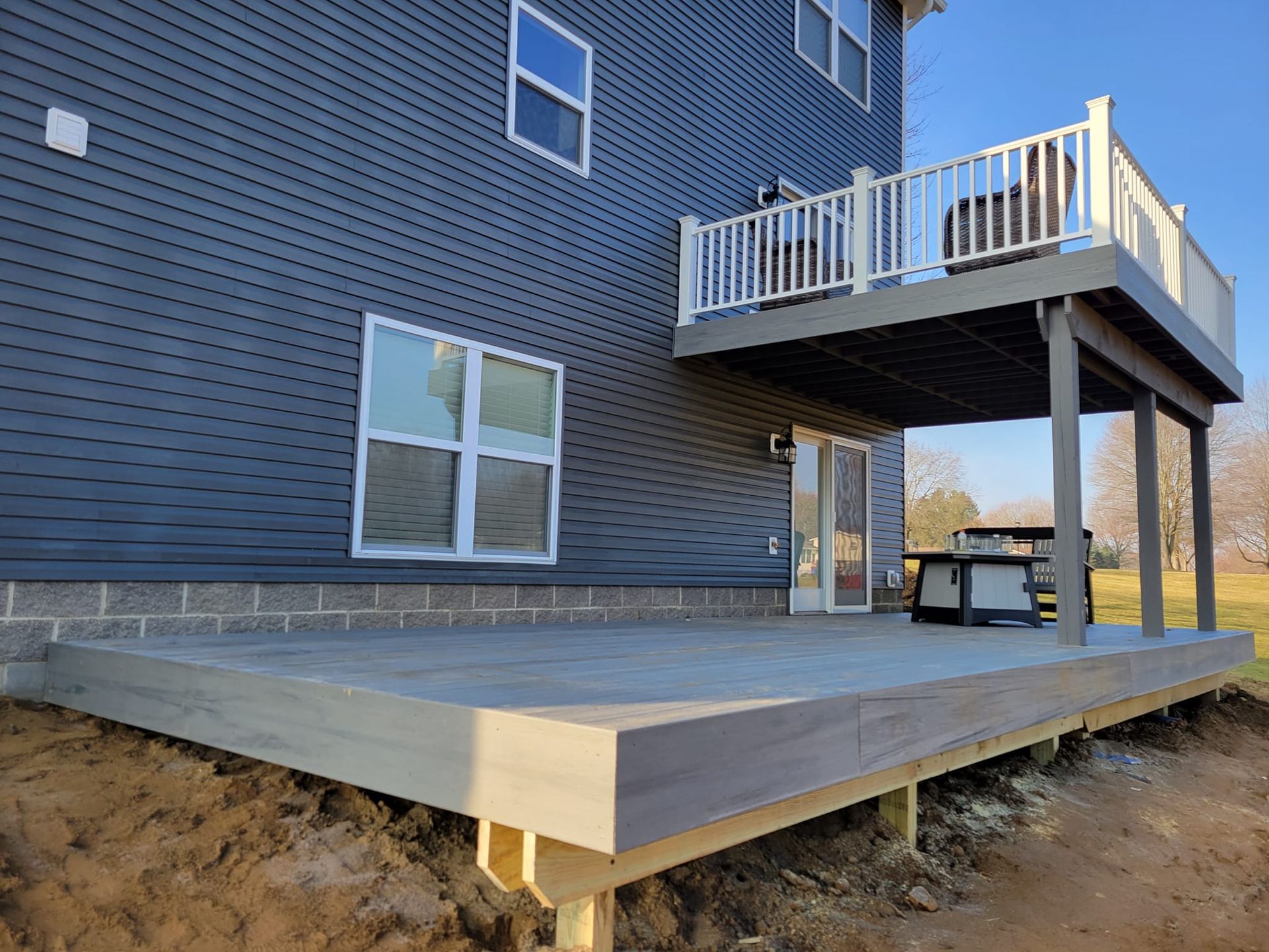 Back of a house with a gray deck.  A second story deck has white railing.  The deck below has a table and chairs.