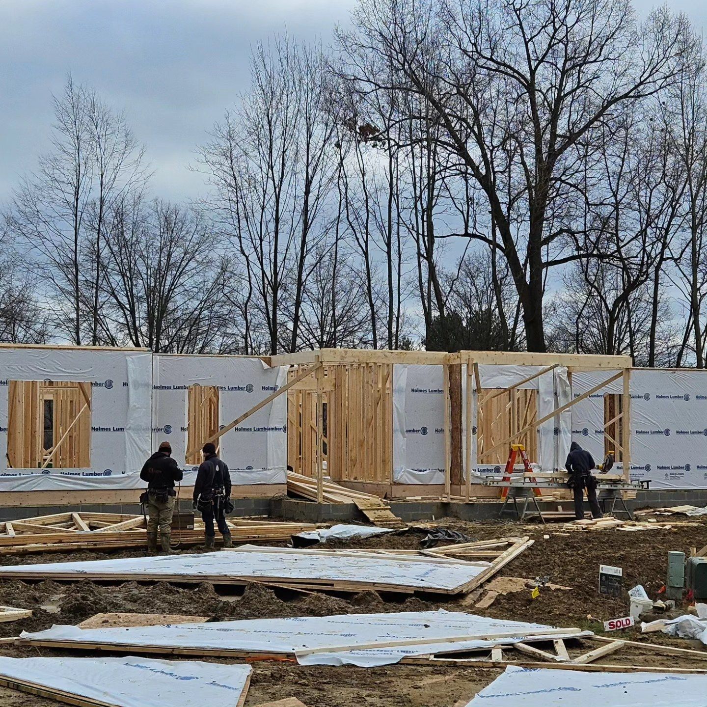 Construction workers framing a building. Wood framing with plastic wrap. Trees in background.