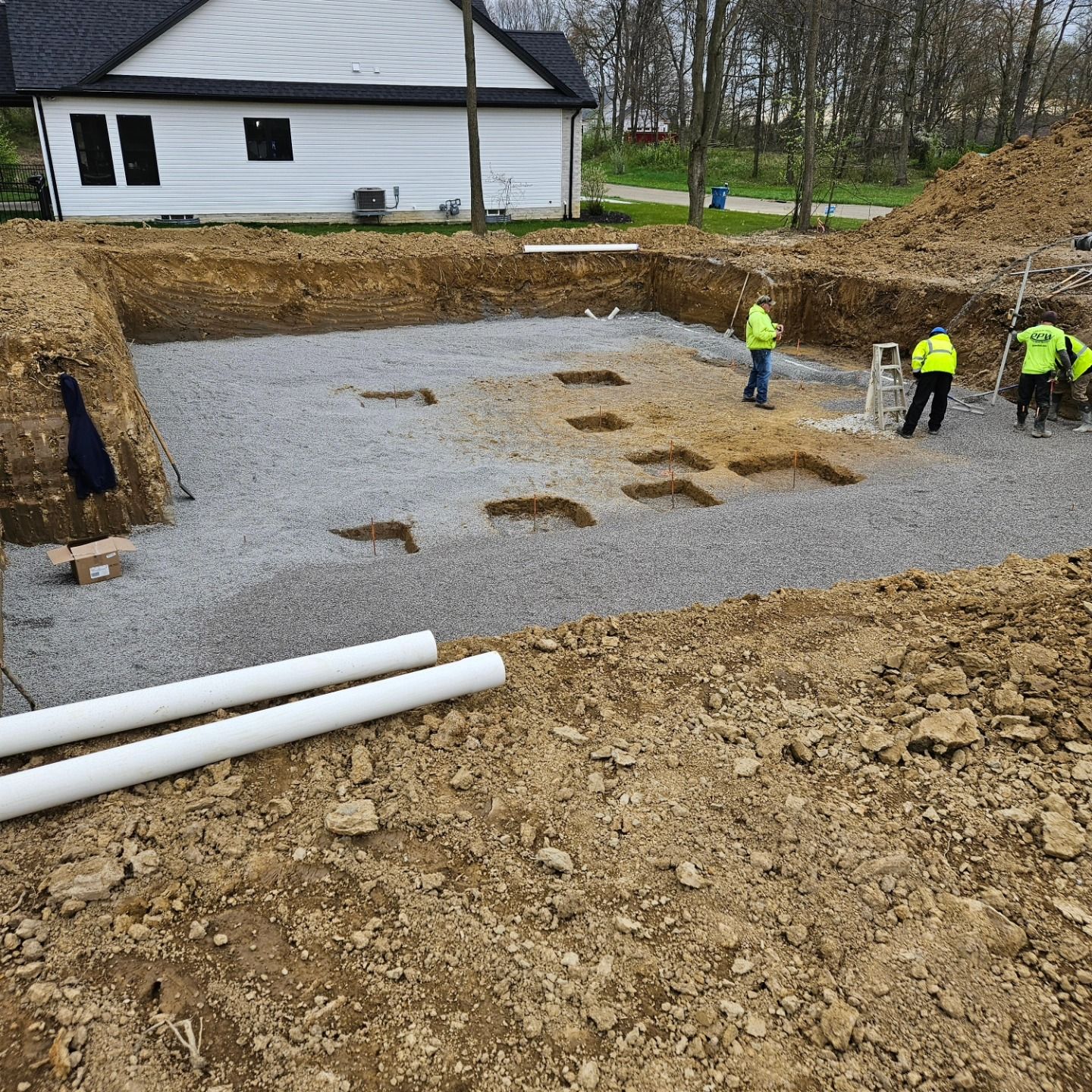 Construction site: excavated foundation with gravel, workers in safety vests. White pipes in foreground, residential house in background.