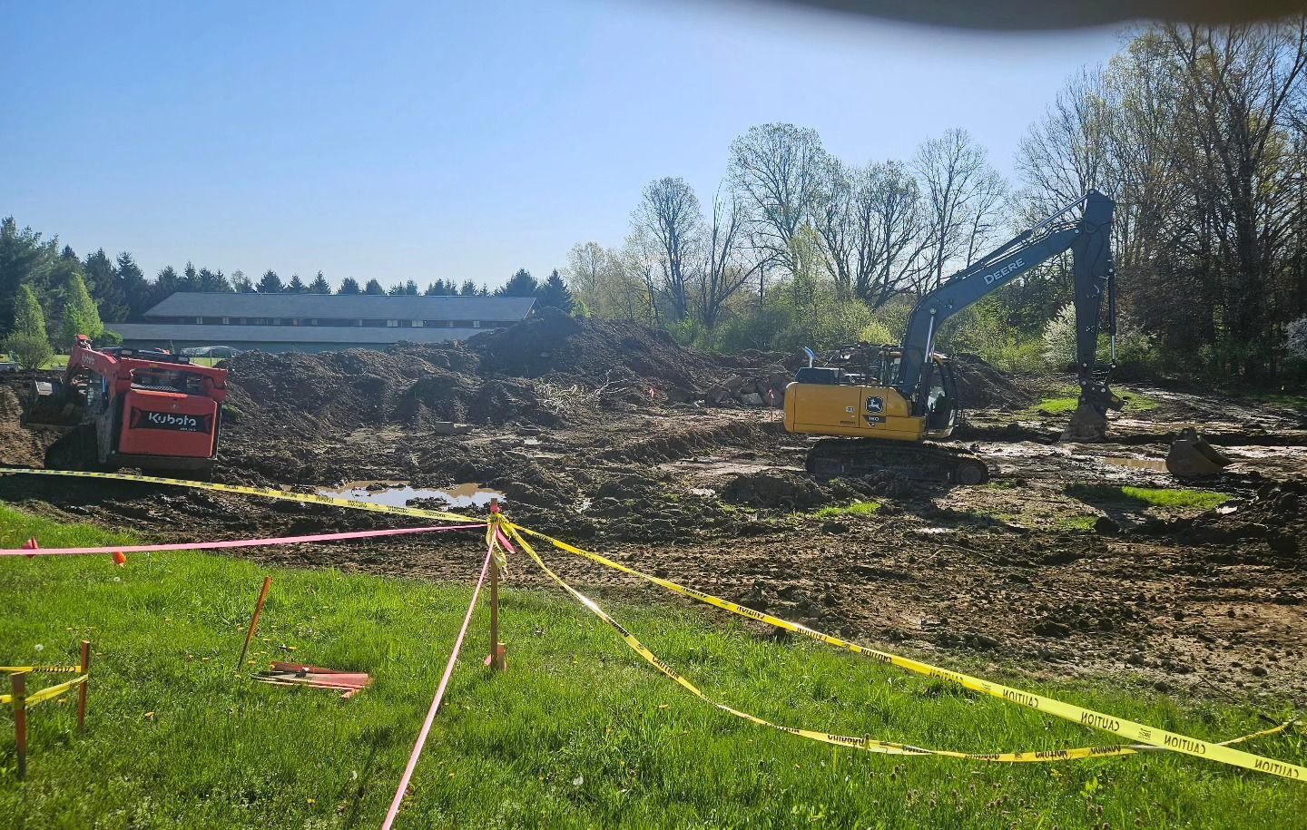 Construction site with excavator and skid steer on muddy ground. Yellow caution tape in foreground. Trees and building in background.