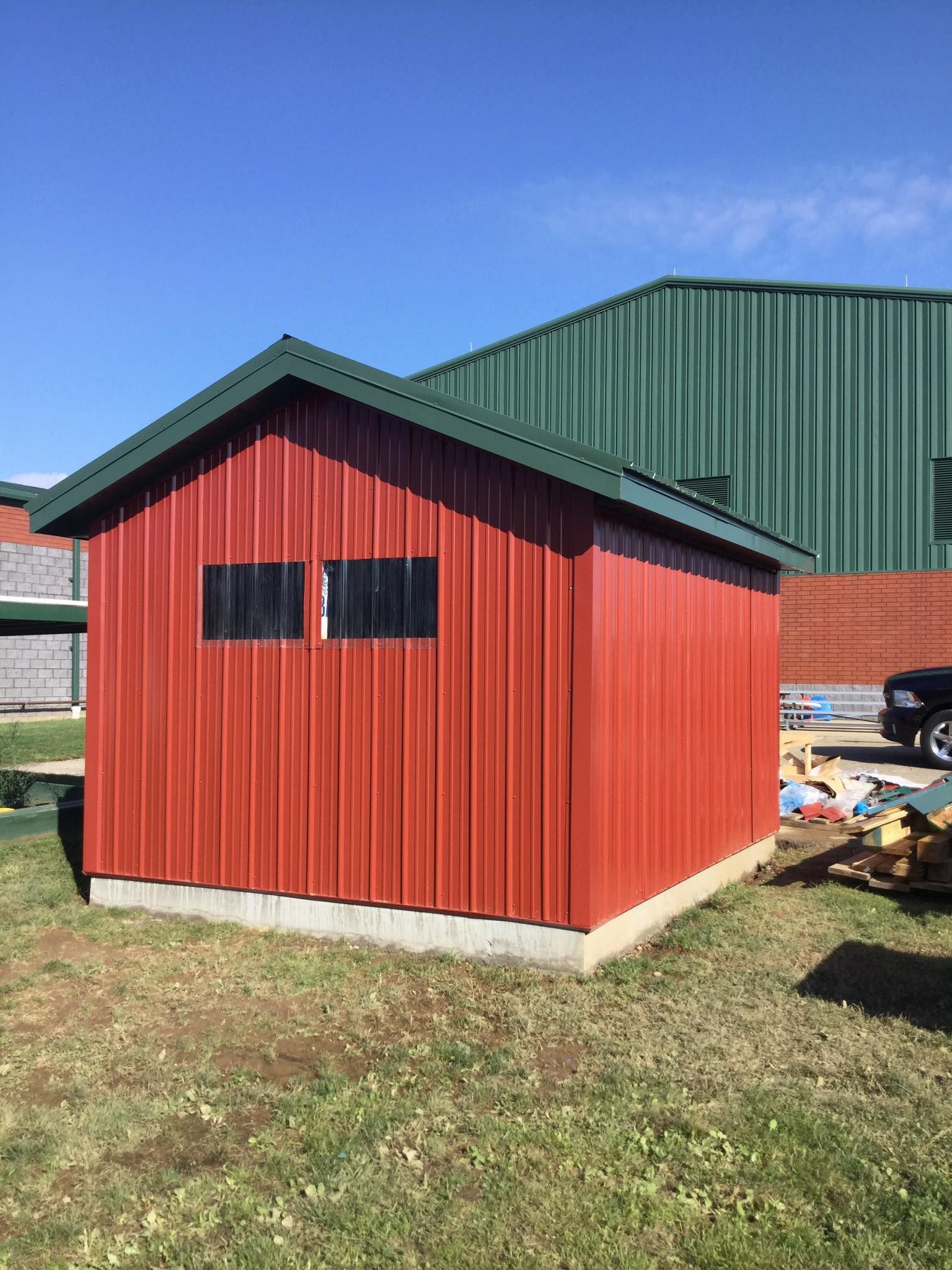 Red metal storage shed with green roof, two small windows, on grass, under blue sky.