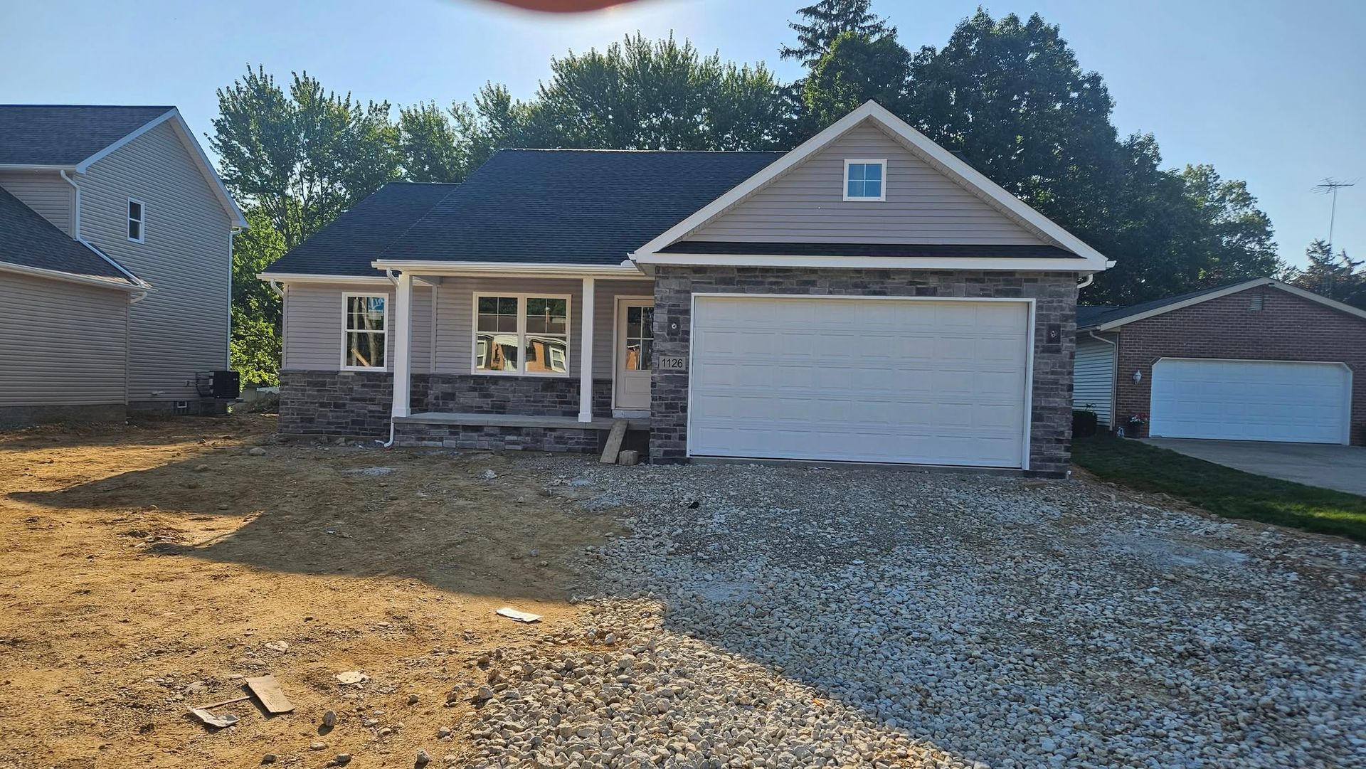 New single-story house with a stone and tan exterior, gray roof, and attached garage, in a dirt and gravel lot.