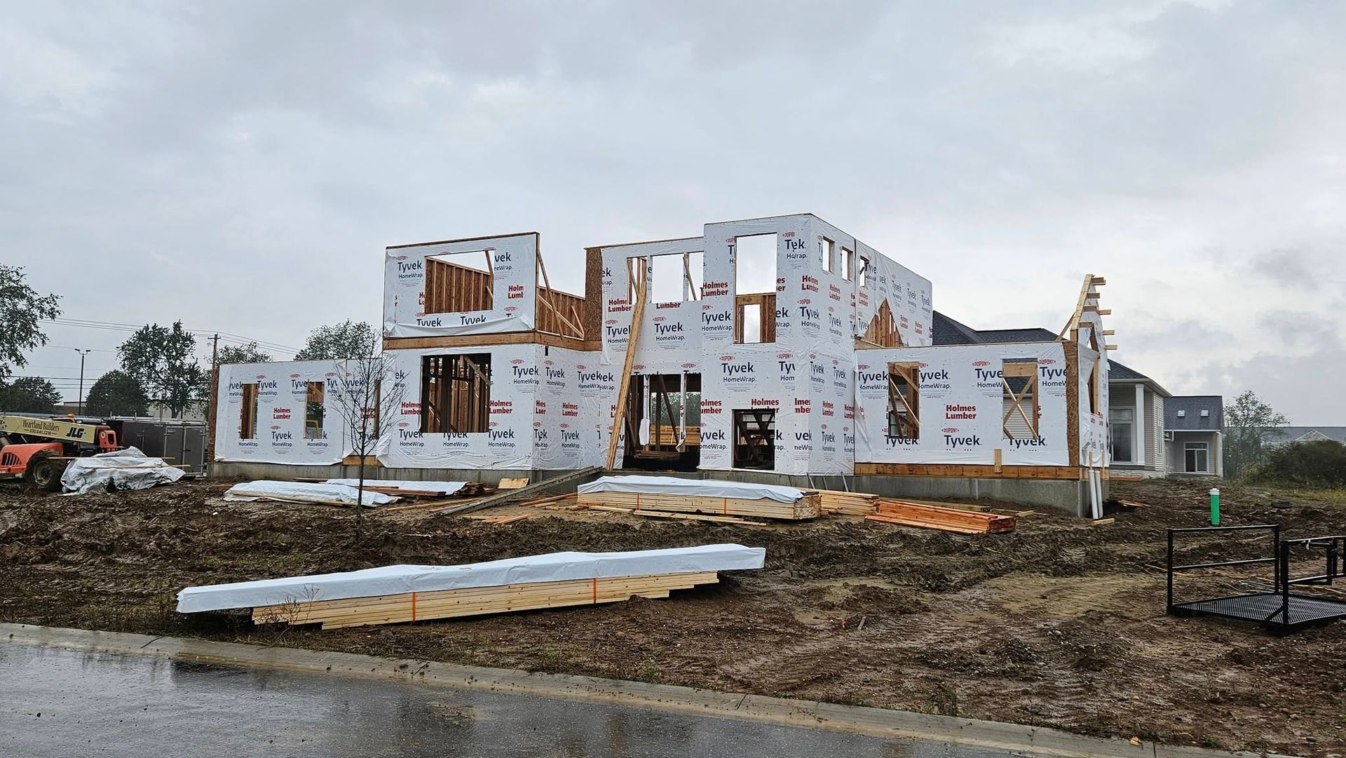House under construction; wood framing wrapped in white sheathing. Dirt, lumber, and heavy machinery around the building. Overcast sky.
