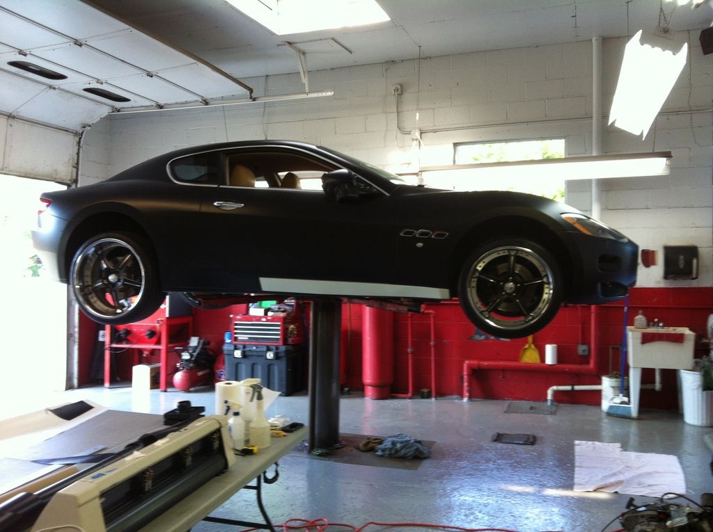 Black Maserati sports car on a lift in a garage with red walls and a cutting machine.