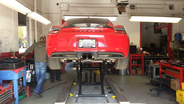Red Porsche sports car on a lift in a garage; two people nearby.