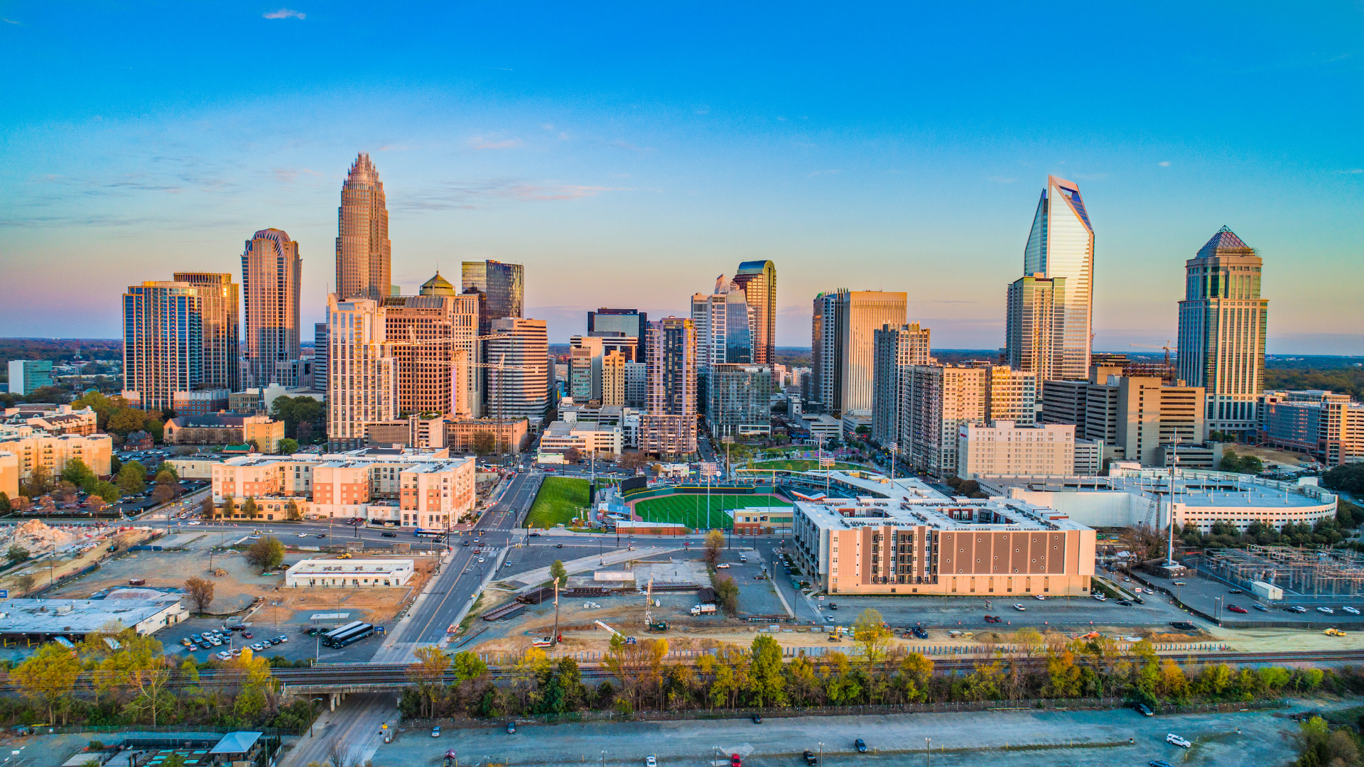 City skyline at sunset with modern high-rises, a prominent tall tower, and a wide boulevard below.