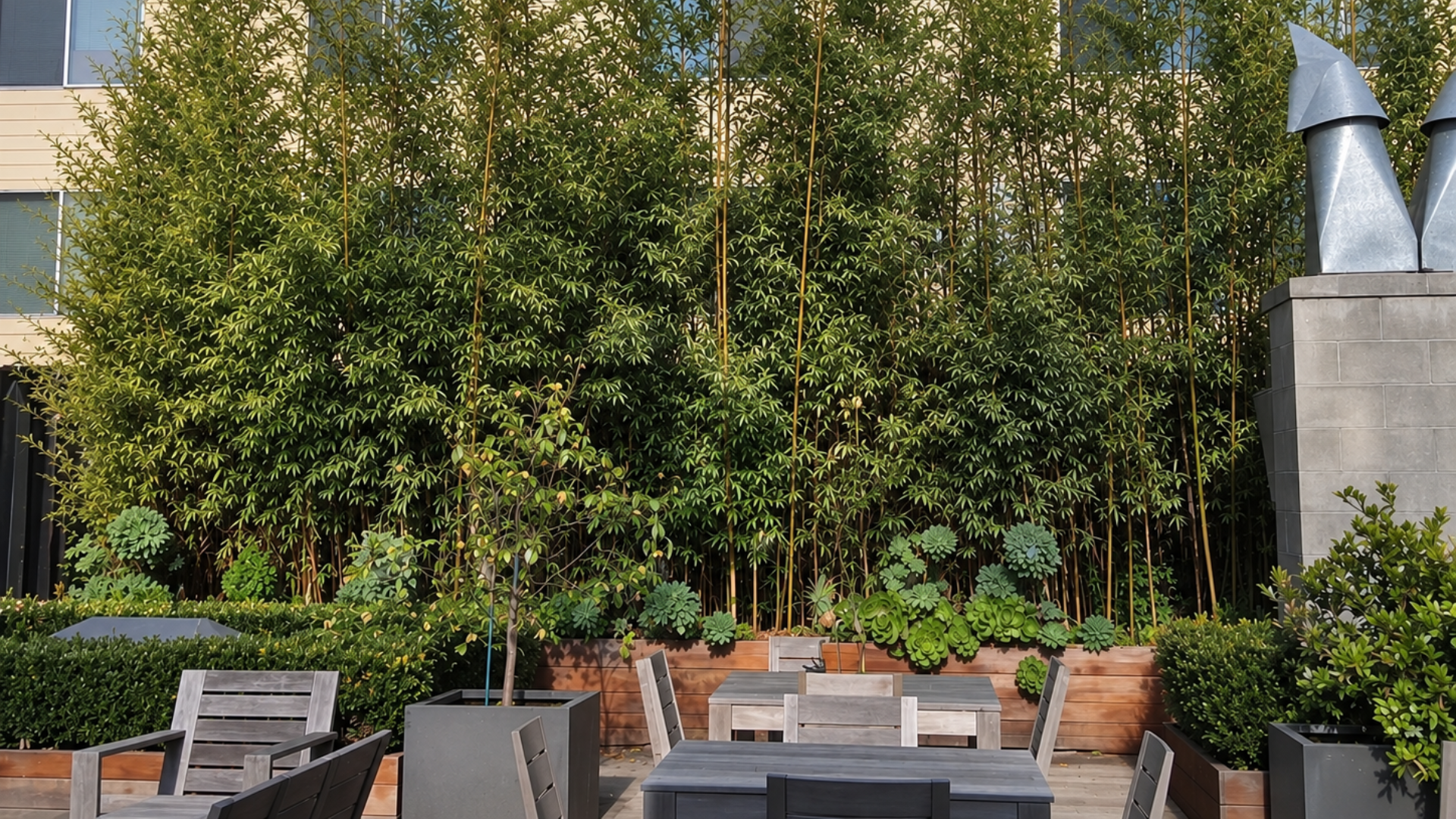 Outdoor patio with gray chairs, wooden tables, and lush green trees and planters,  a work of Jungle Bamboo and Palm Nursery