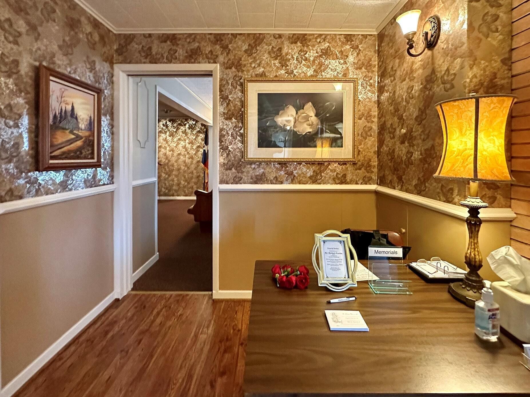 Hallway with floral wallpaper, wood floor, desk lamp, and framed paintings in a warmly lit home interior