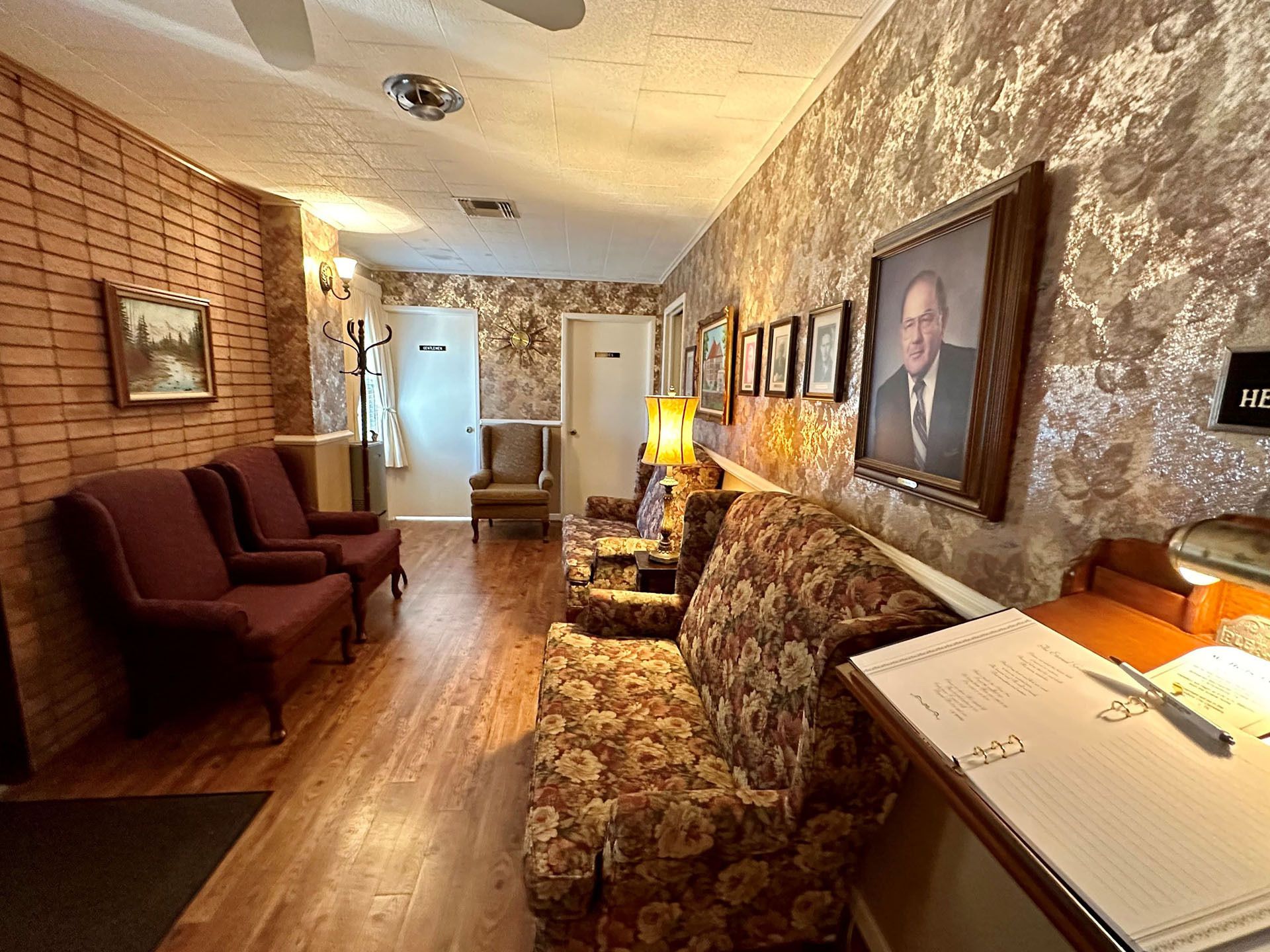 Narrow hallway with patterned chairs, framed portraits, brick walls, and warm lamp lighting