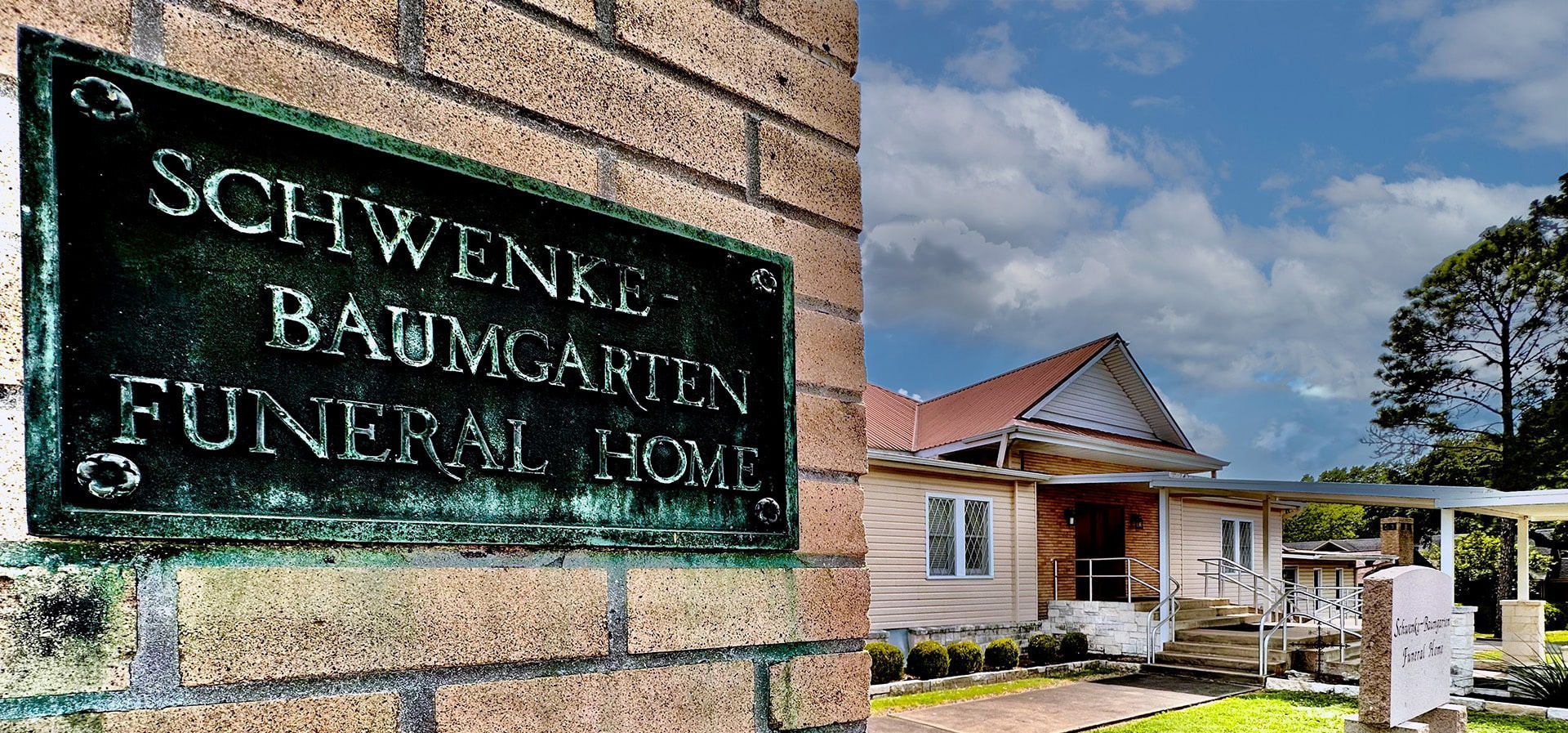 Schwenke-Baumgarten Funeral Home sign on a brick building beside a small house and driveway