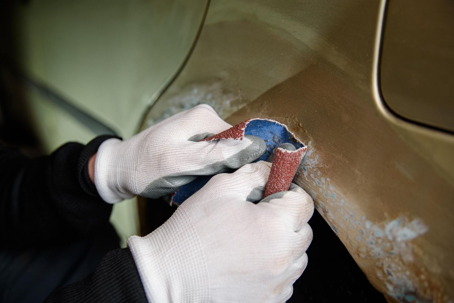 Gloved hands sanding a brown car panel with sandpaper during bodywork
