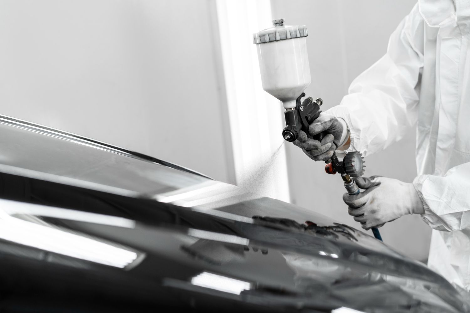 Person spray-painting a car hood in a workshop, using a spray gun with white paint.