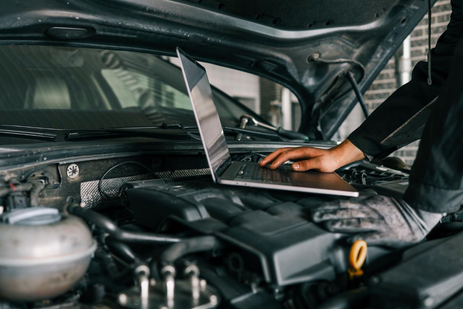Mechanic working on a car engine with the hood open, using a laptop in the engine bay