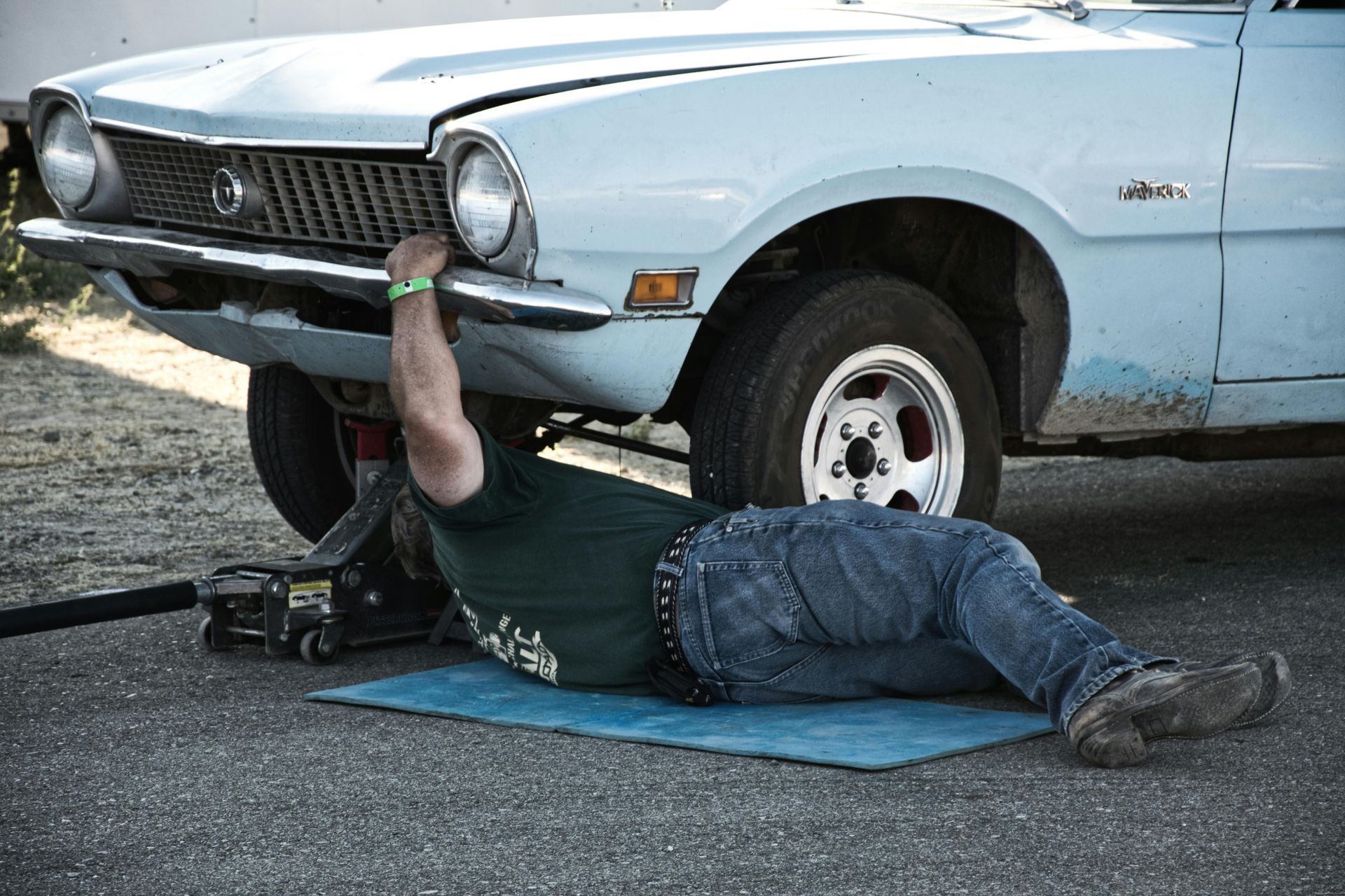 Mechanic working under a light blue car on a jack, lying on a blue mat.