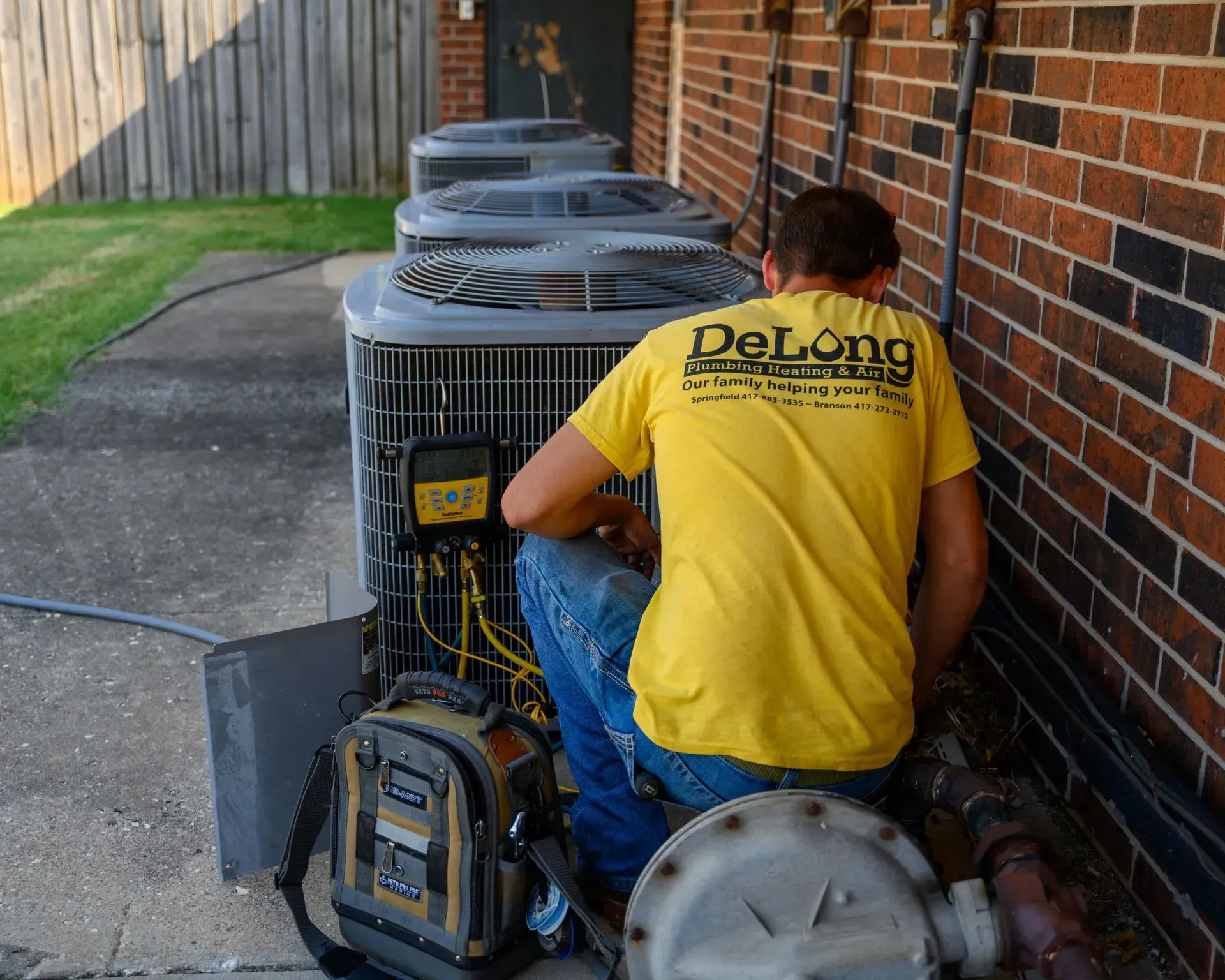 Technician in yellow shirt servicing outdoor HVAC units beside a brick wall.