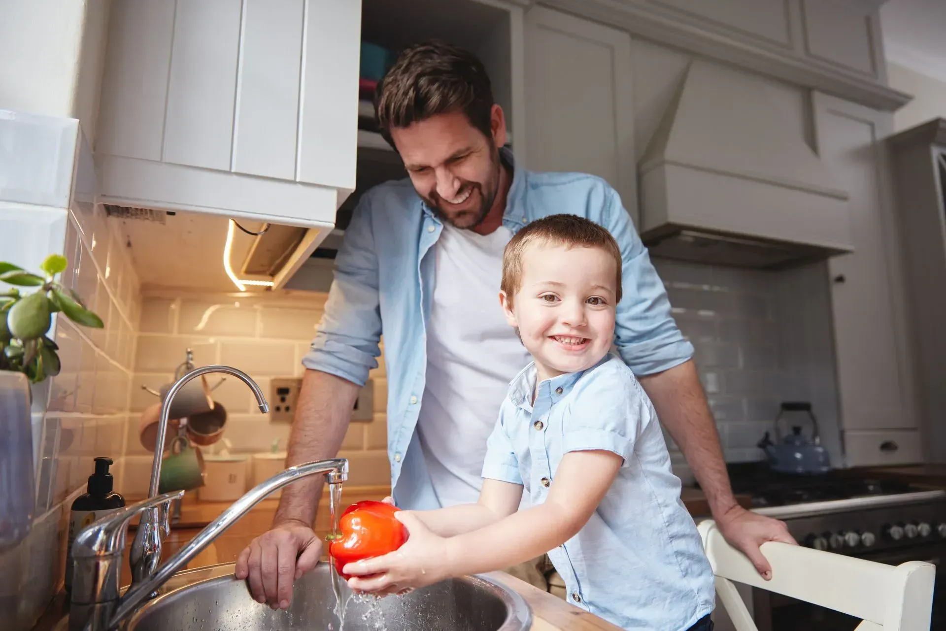 Man and child smiling in a bright kitchen, washing vegetables at the sink.