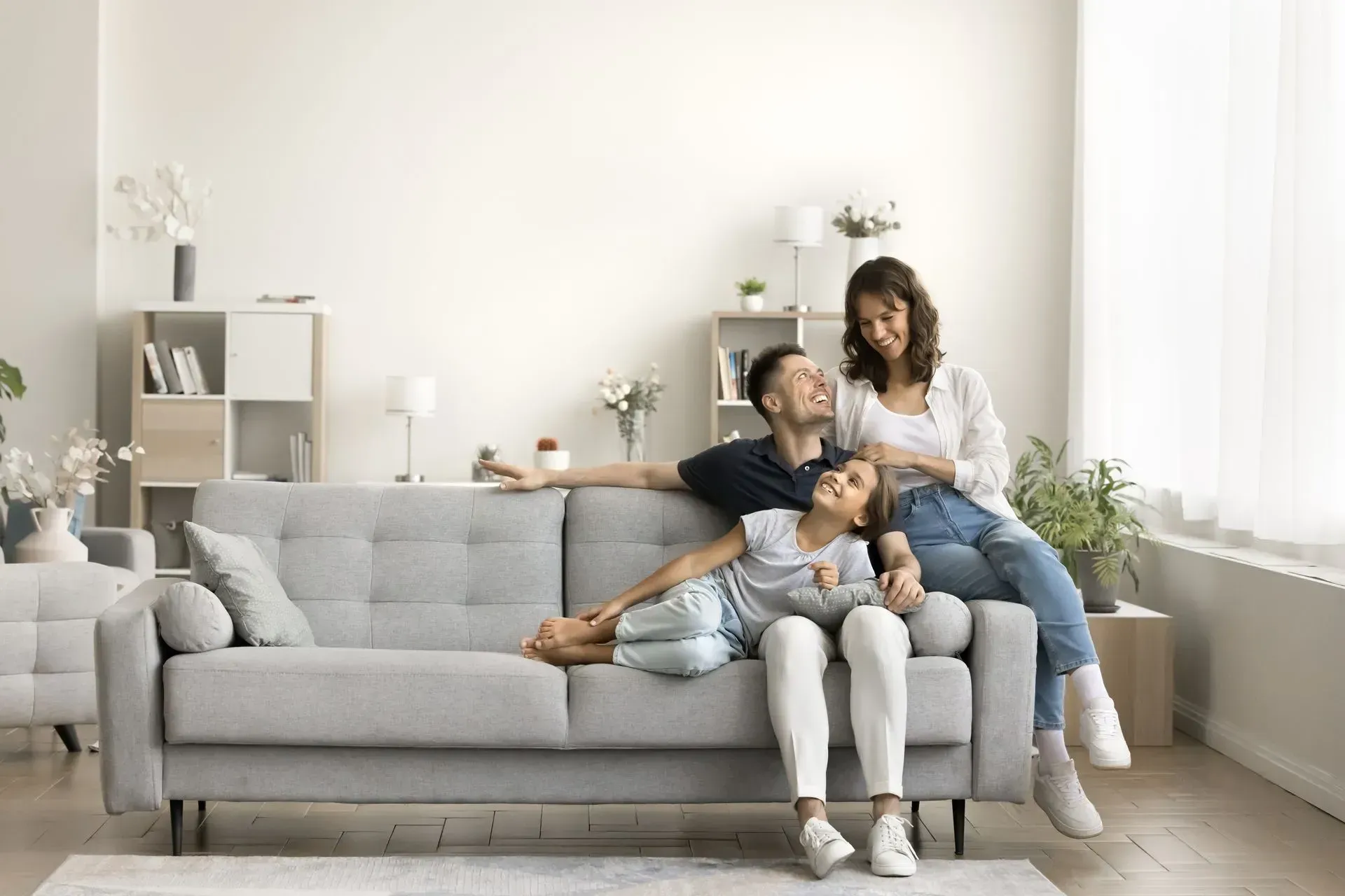 Family sitting on a light gray sofa in a bright living room with white decor and sunlight.