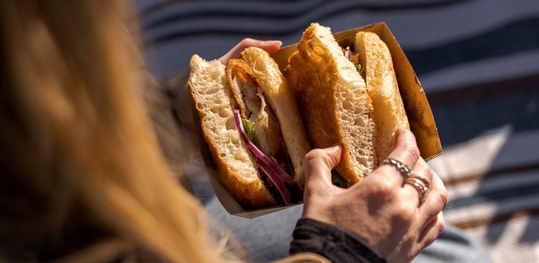 Looking over the shoulder of a women as she picks up a sandwich sitting on a blue and brown stripped picnic blanket