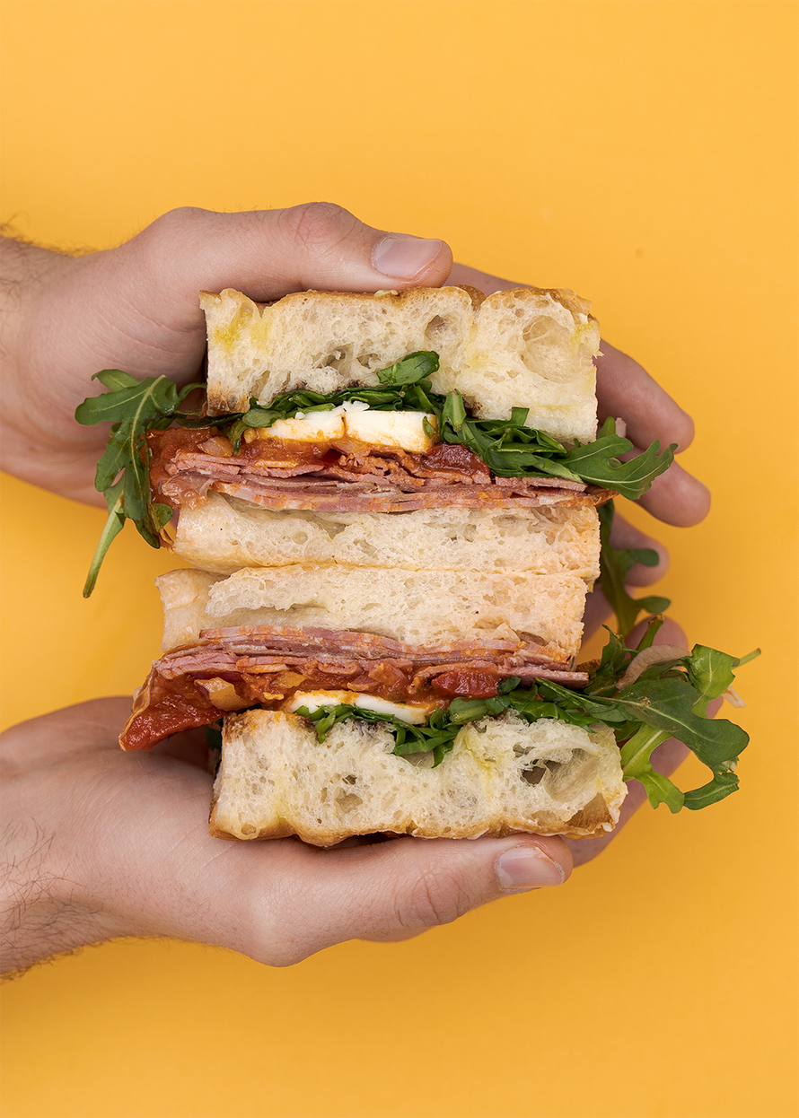 Hands holding the Triple Threat Sandwich on a mustard yellow background
