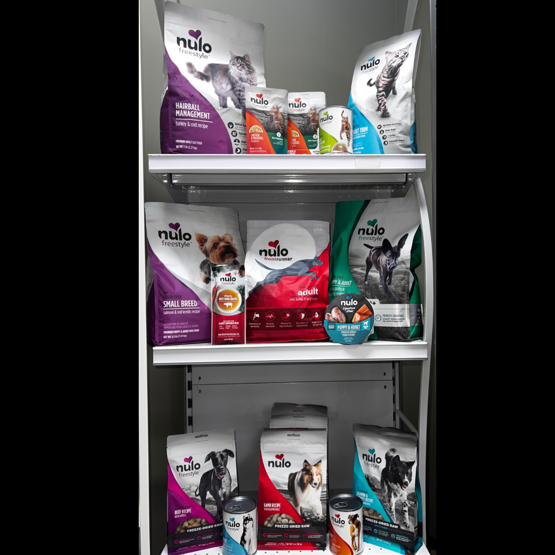 Pet food bags displayed on store shelves, mostly white with purple, red, teal, and black packaging