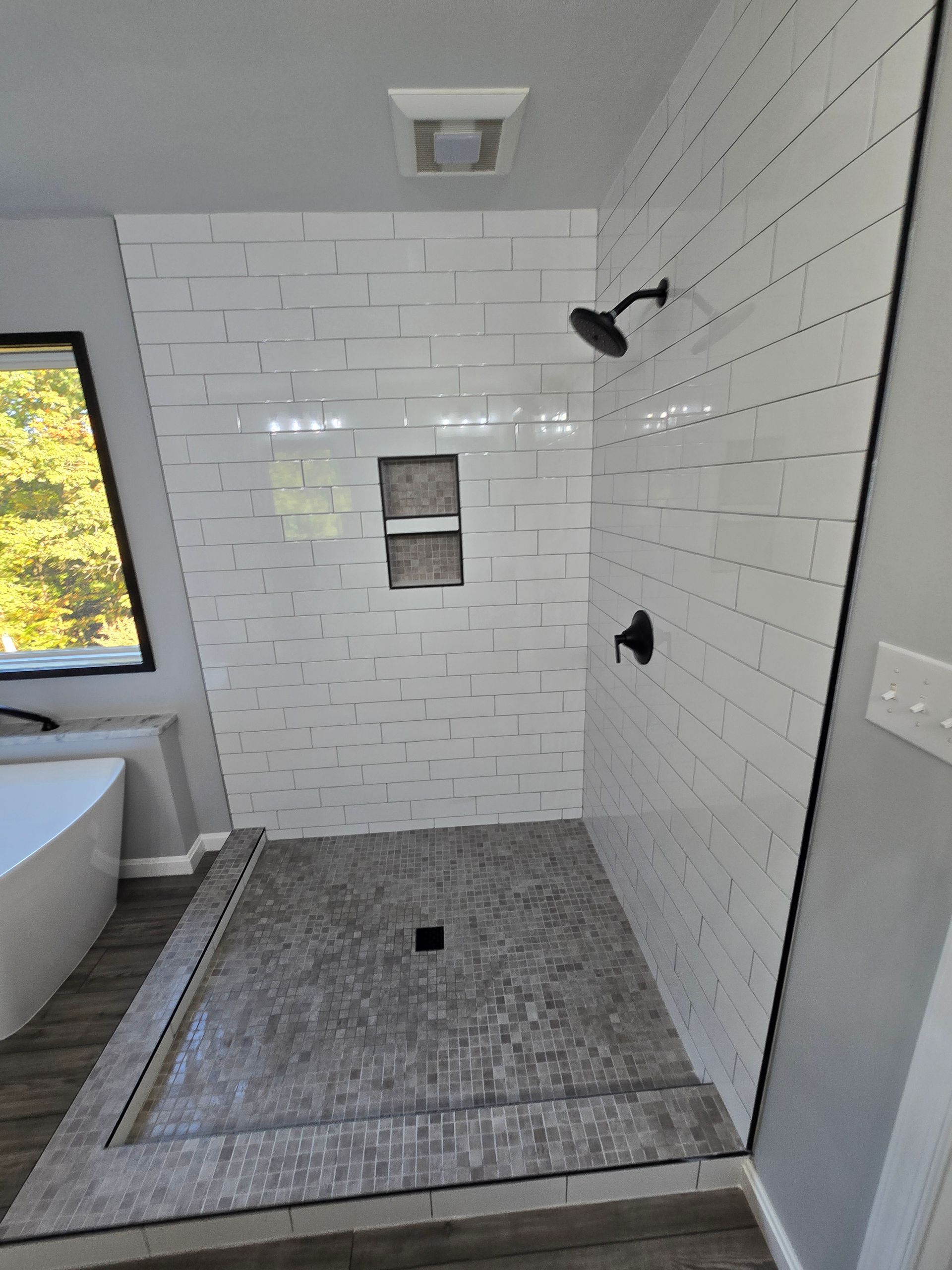 Bathroom with white tile shower, dark fixtures, and a gray stone floor. White walls.