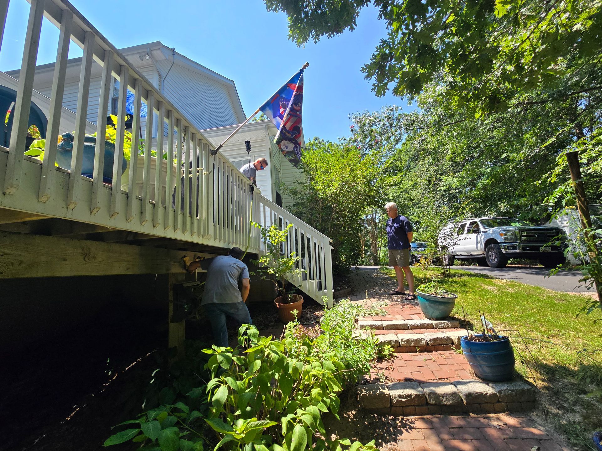 People working on a house with a deck. A flag flies, a vehicle is parked nearby.