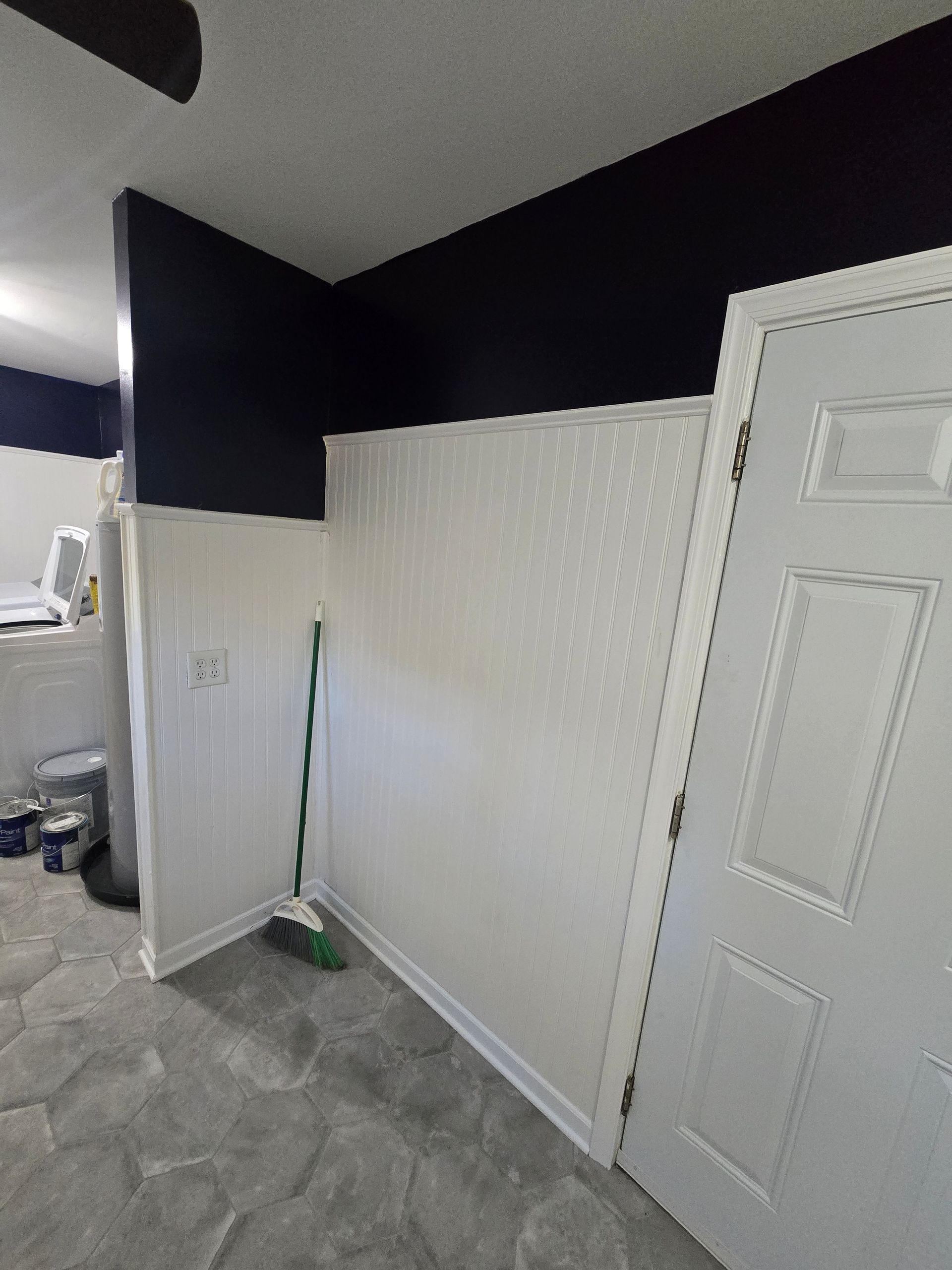 A room with white paneling and a navy blue upper wall. A broom rests against the wall. A white door is on the right.