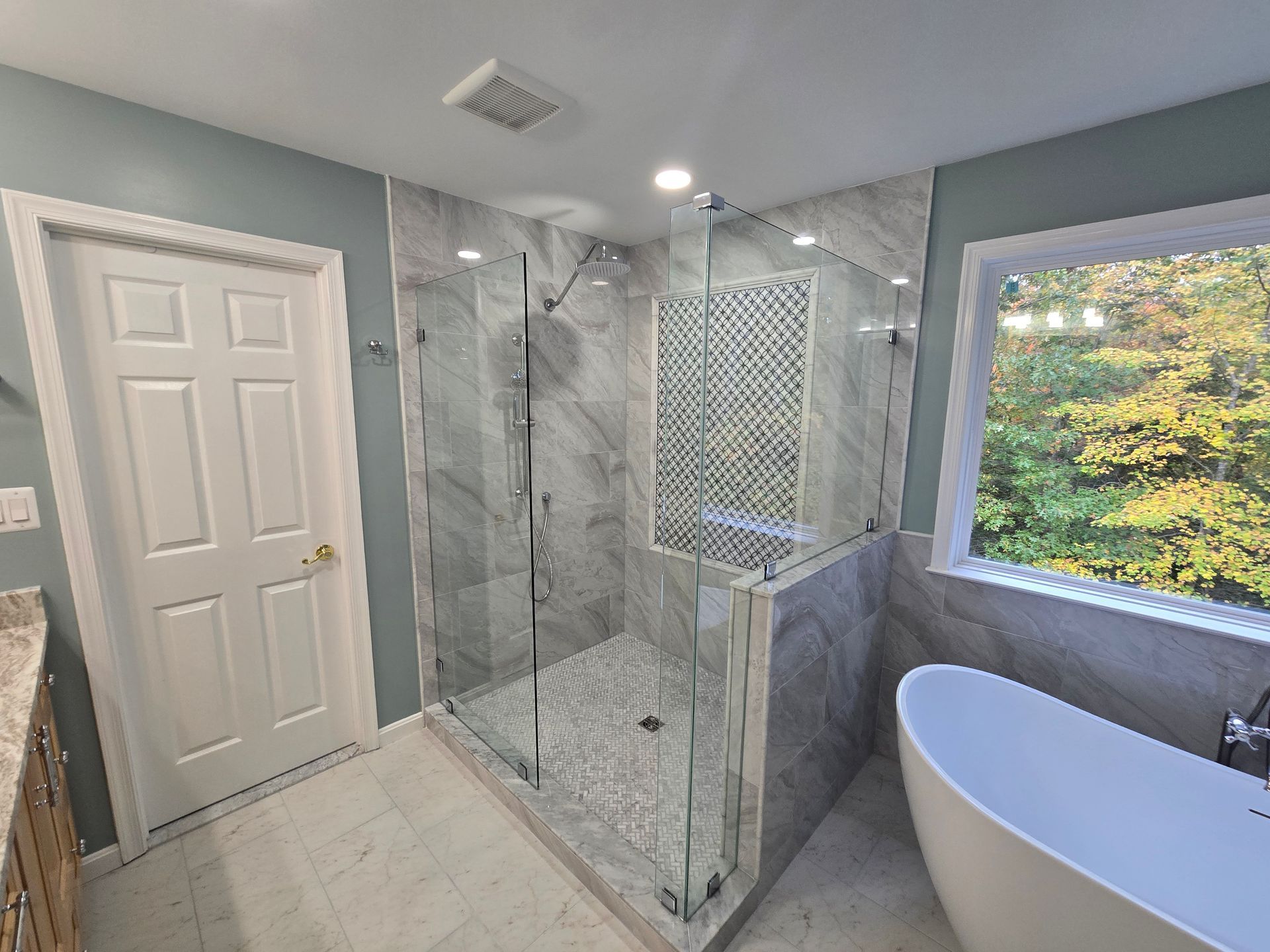 Bathroom with a large glass shower, a white bathtub, and a window overlooking greenery.