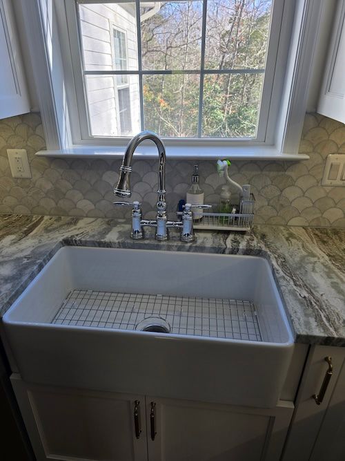 White farmhouse sink under a window in a bright kitchen with marble countertops and chrome faucet.
