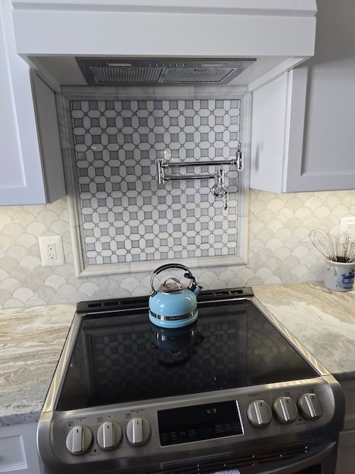 Kitchen stove with a blue kettle beneath a patterned tile backsplash and range hood