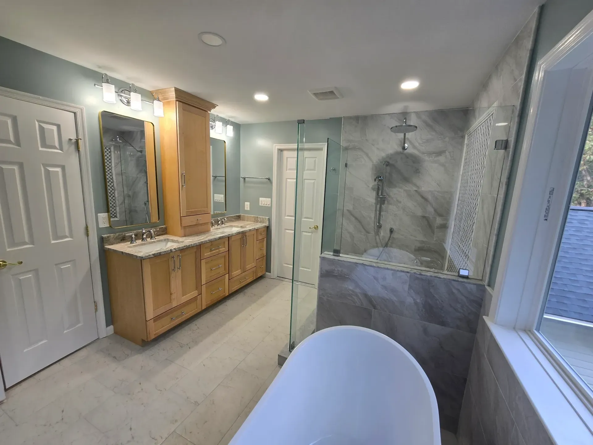 Bathroom with light wood cabinets, gray tiled shower, and a white soaking tub.