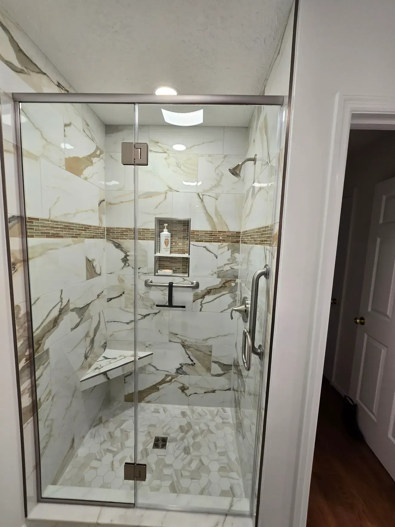 Modern shower with marble-like tiles, glass door, and chrome fixtures. Includes a built-in bench and soap niche.