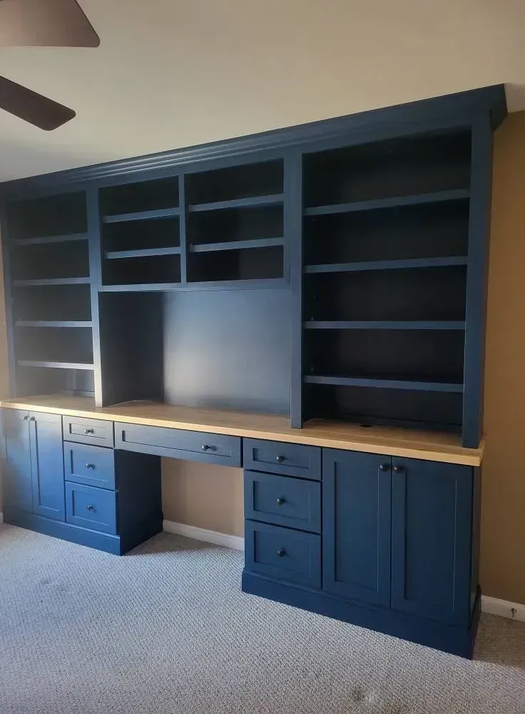 Built-in dark blue desk with light wood top, shelves, and cabinets against a tan wall.