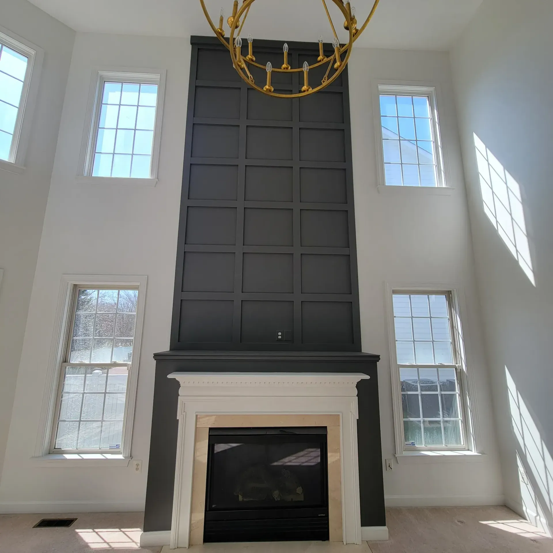 Living room with fireplace, gray panel wall, gold chandelier, and multiple windows.