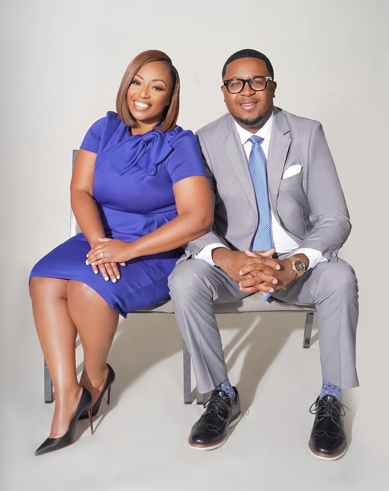 Two people seated on chairs, smiling at the camera, wearing blue and gray business attire.
