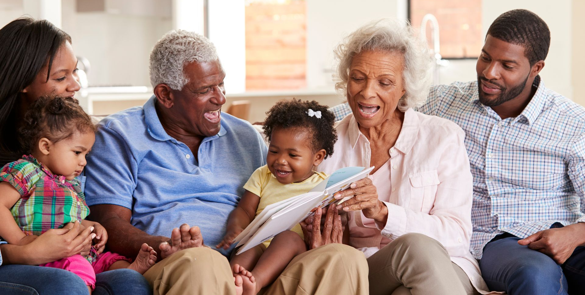 Three generations sitting together on a couch, smiling and reading a book with a child.